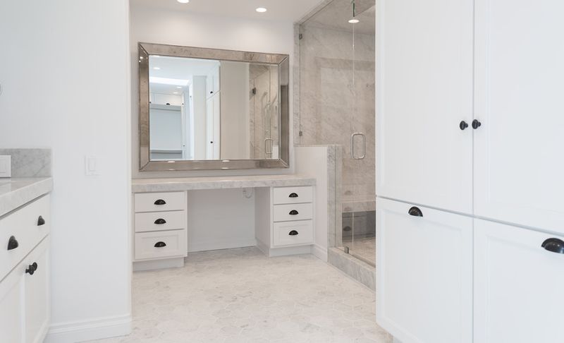 A bright, elegant master bathroom featuring white cabinetry with black hardware, a marble-topped makeup vanity, large silver-framed mirror, and a glass-enclosed marble tile shower.