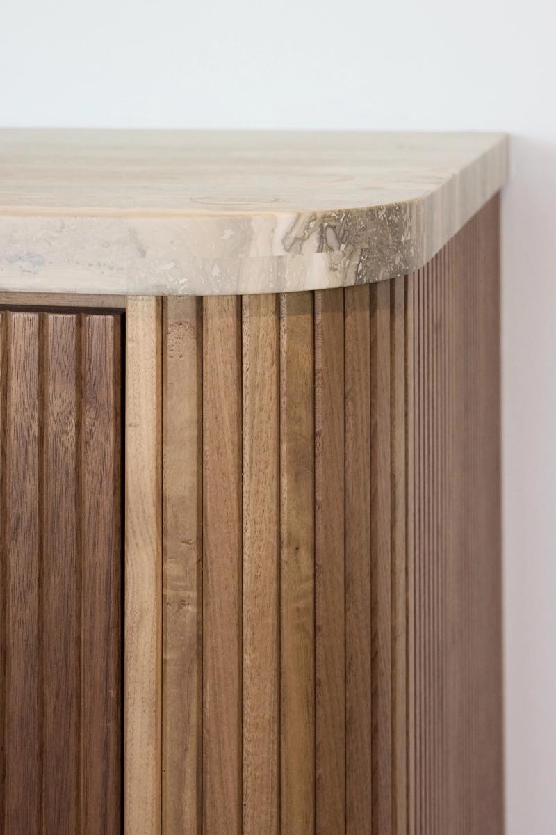 A close-up detail shot of a custom cabinet featuring vertical fluted wood panels in a warm walnut tone, topped with a curved travertine or fossil stone surface with natural veining.