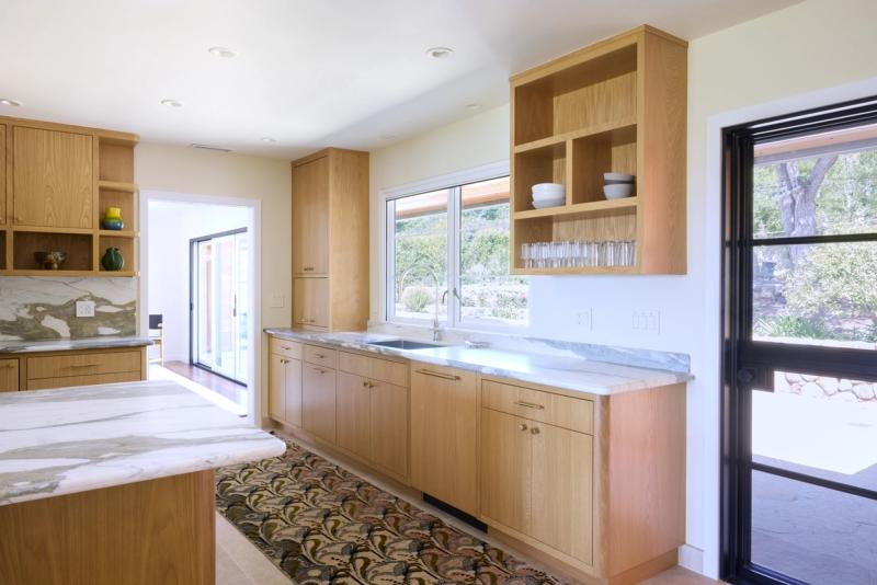 A bright, modern kitchen featuring custom white oak cabinetry with flat-panel doors, marble countertops, open upper shelving, and a window overlooking a garden. A decorative area rug runs along the center of the tile floor.