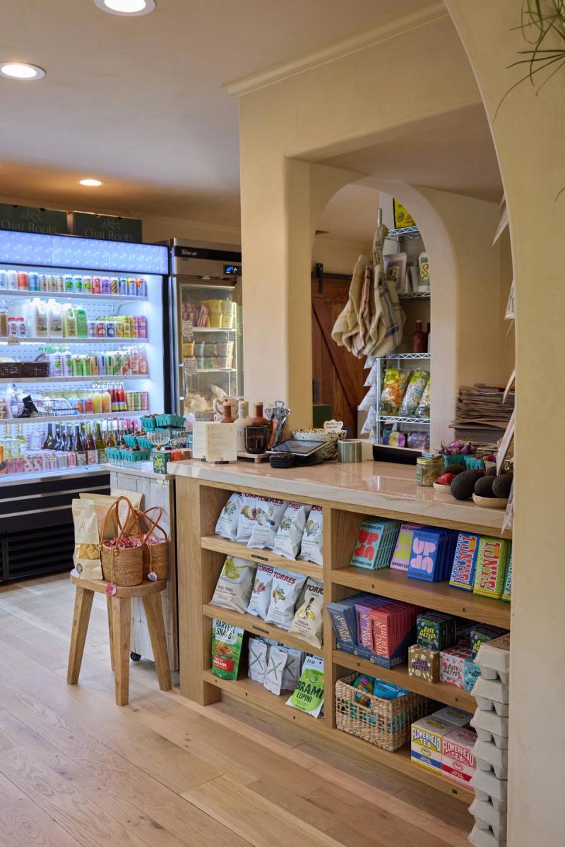 A warm, light-filled specialty grocery or market interior featuring a custom wood checkout counter with open shelving stocked with snacks, teas, and chocolates, alongside a large illuminated refrigerated display case in the background.
