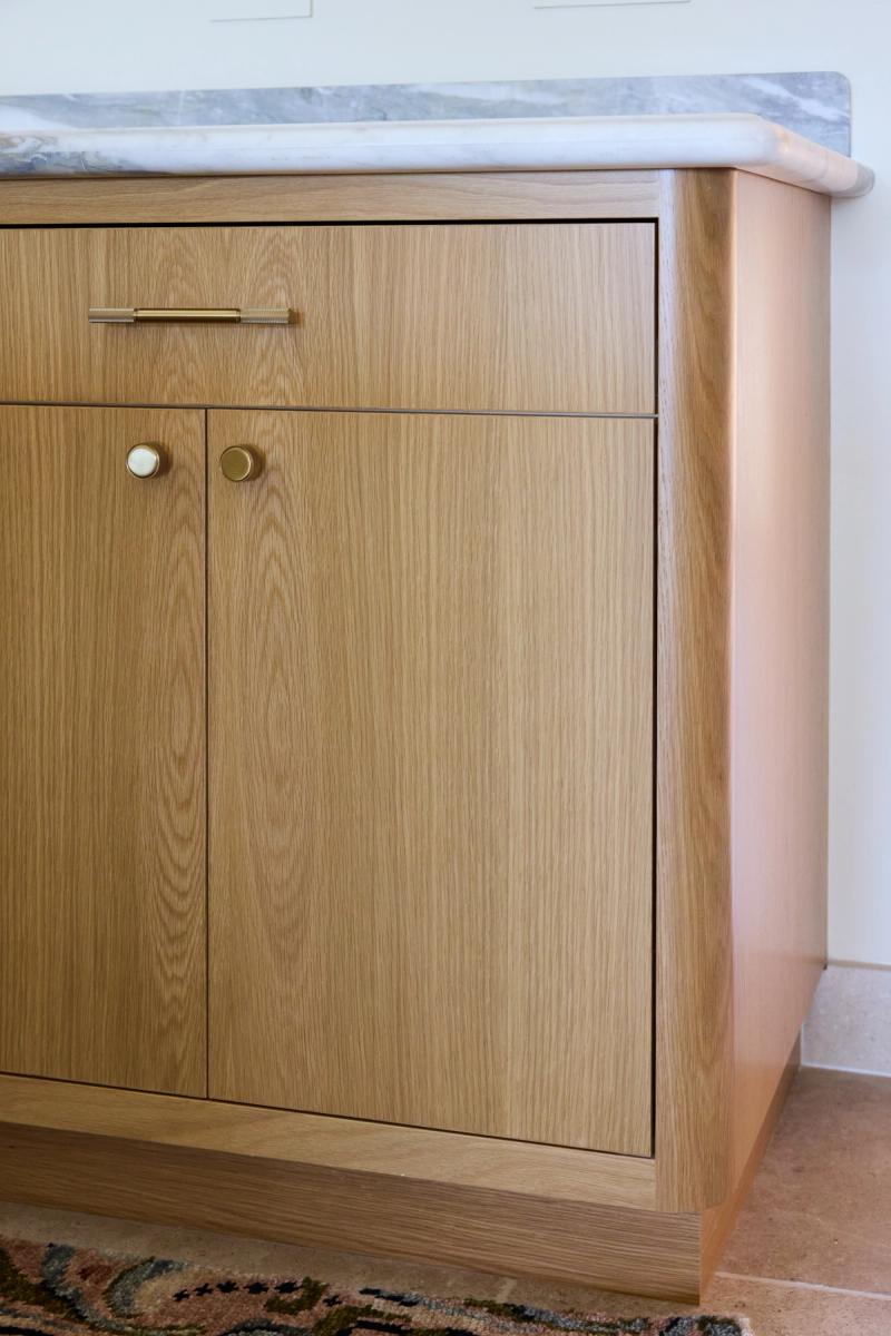 A close-up of a white oak bathroom vanity featuring flat-panel cabinet doors with brass knobs and a bar pull, topped with a gray veined marble countertop.
