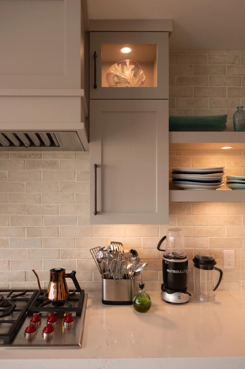A warmly lit kitchen corner featuring gray shaker-style cabinetry with a glass-front display cabinet, cream brick tile backsplash, open shelves with teal glassware and stacked plates, and a white quartz countertop with a gas cooktop, copper kettle, utensil holder, and NutriBullet blender.