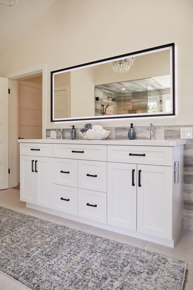 A spacious master bathroom featuring a large white shaker-style double vanity with black hardware, white quartz countertop, gray tile backsplash, and a wide LED-backlit mirror reflecting a walk-in shower.