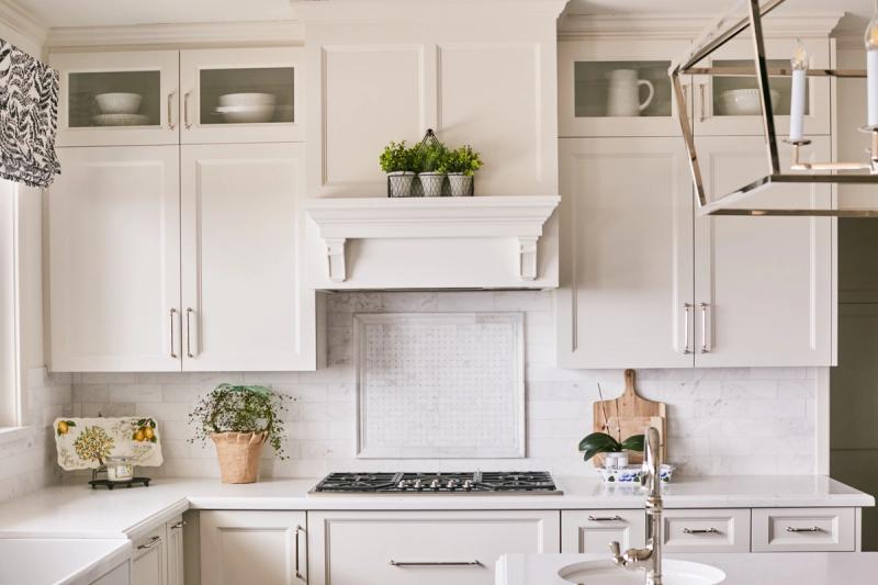 A bright, traditionally styled kitchen featuring custom white cabinetry with raised panel doors, a decorative mantel-style range hood, marble tile backsplash, and glass-front upper cabinets displaying white dishware.