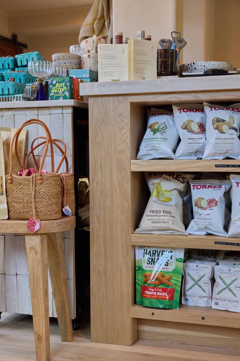 A light natural oak open shelving unit stocked with gourmet snack products including Torres chips and Harvest Snaps, displayed in a warm boutique retail setting alongside woven baskets and decorative items.