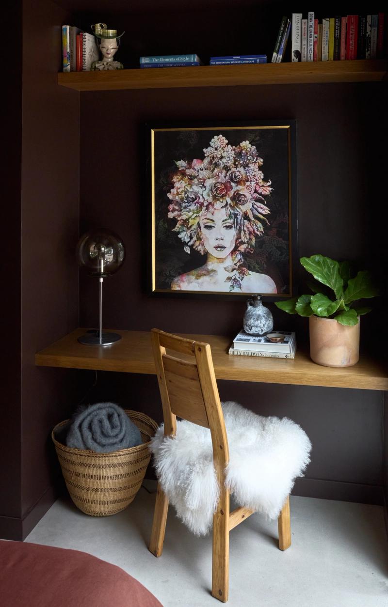 A cozy built-in home office nook featuring a floating oak desk and overhead shelf, set against deep brown walls, styled with books, a globe lamp, potted plant, and a floral portrait painting.