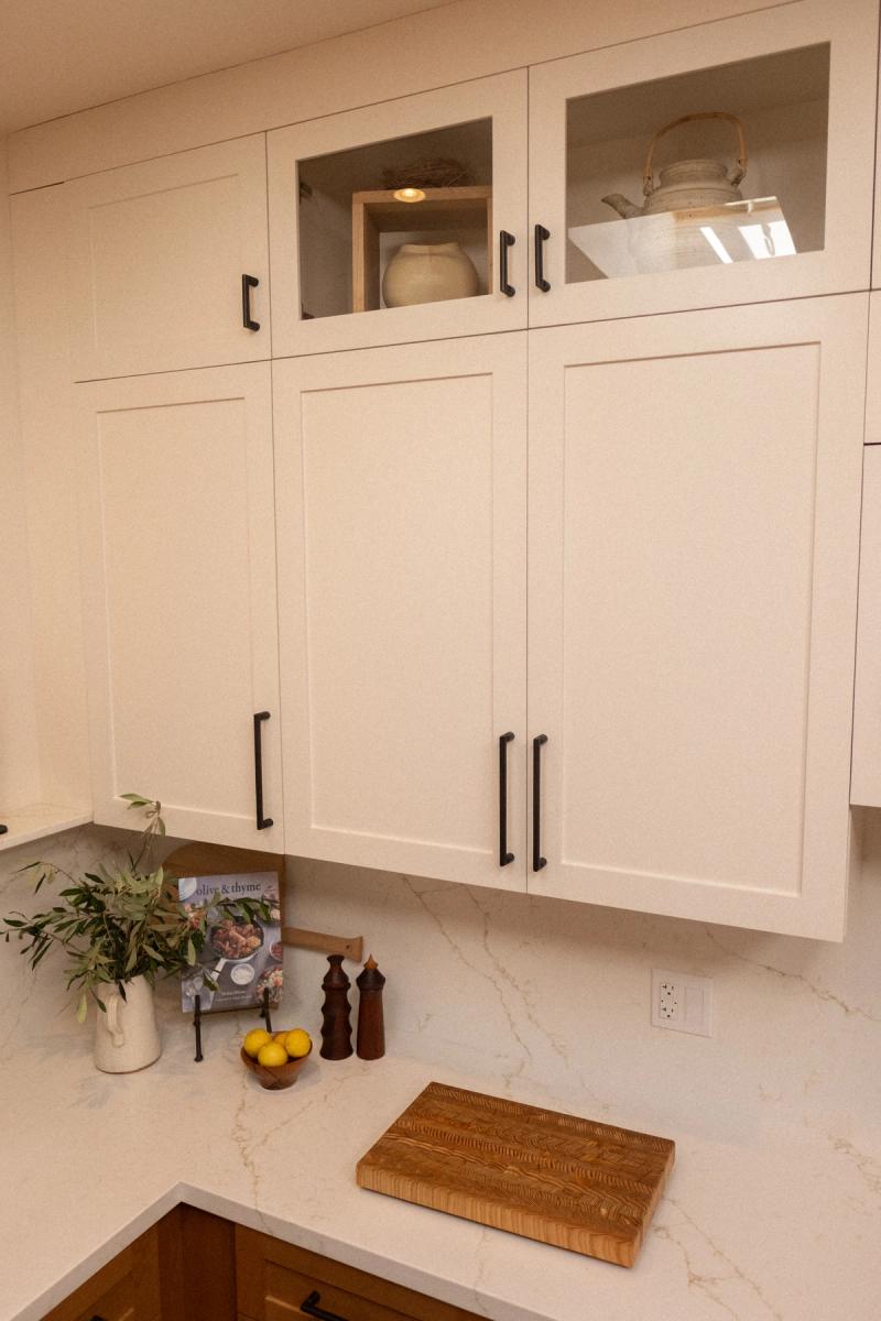 Cream white shaker-style upper kitchen cabinets with matte black hardware, glass-front display cabinets at top, and a white quartz countertop styled with lemons, a cookbook, salt and pepper mills, and a wood cutting board.