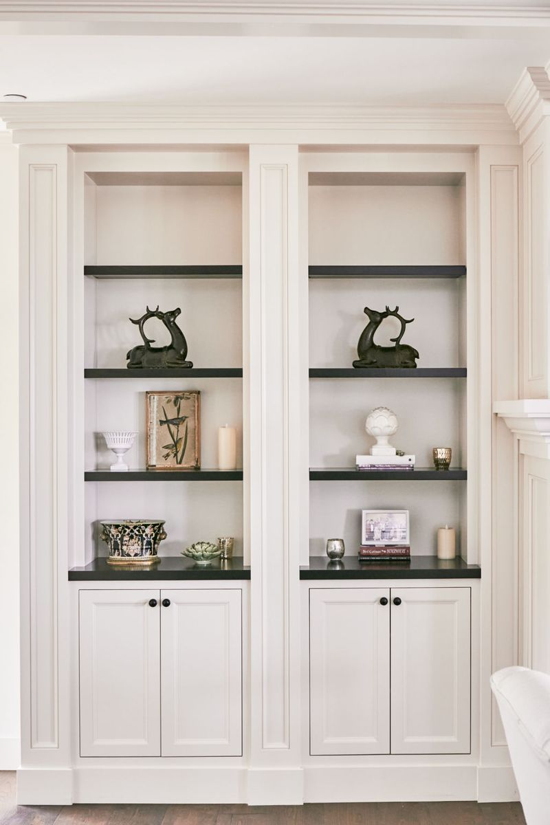 A pair of elegant cream-painted built-in bookcases with dark shelves, styled with decorative objects including deer figurines, botanical art, and ceramic accents, above shaker-style lower cabinet doors with black hardware.