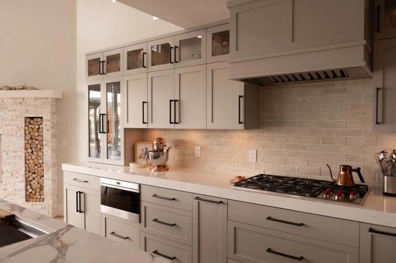A custom kitchen featuring greige shaker-style cabinetry with matte black hardware, white quartz countertops, a travertine subway tile backsplash, stainless steel gas cooktop, and a built-in oven. A stone fireplace with stacked wood is visible to the left.