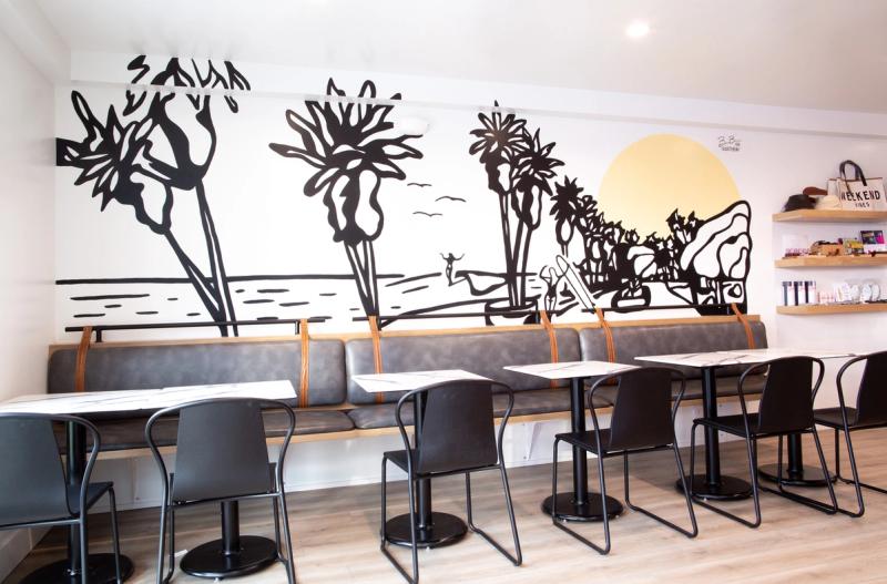 A bright cafe or restaurant interior featuring a long gray leather banquette with wood trim along a white wall painted with a black-and-white beach mural. Several black chairs and marble-top tables are arranged in front of the seating.