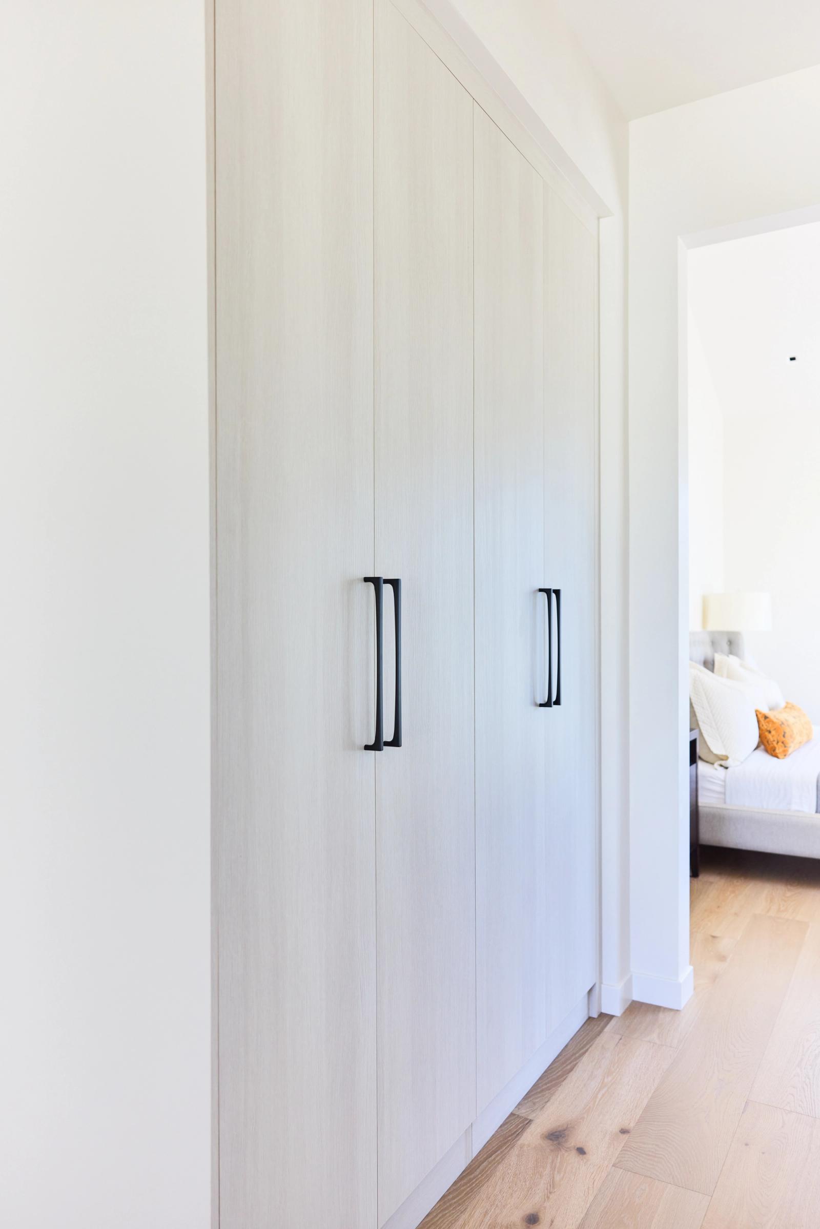 A floor-to-ceiling built-in wardrobe with light white oak panel doors and sleek matte black vertical pull handles, situated in a hallway leading to a bright bedroom.