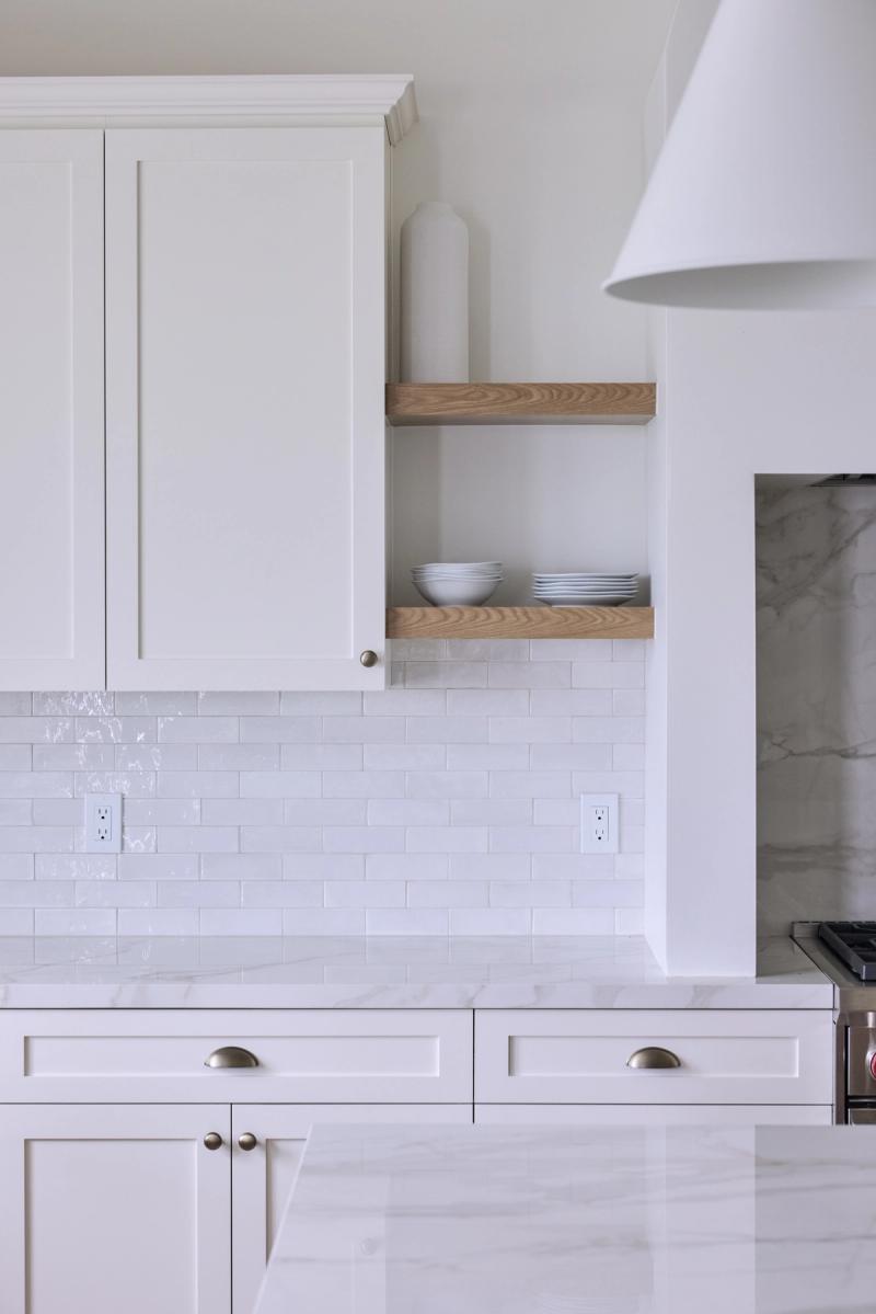 A bright, all-white custom kitchen featuring shaker-style cabinetry, marble countertops, white subway tile backsplash, brushed nickel hardware, and floating oak open shelves styled with white dishes.
