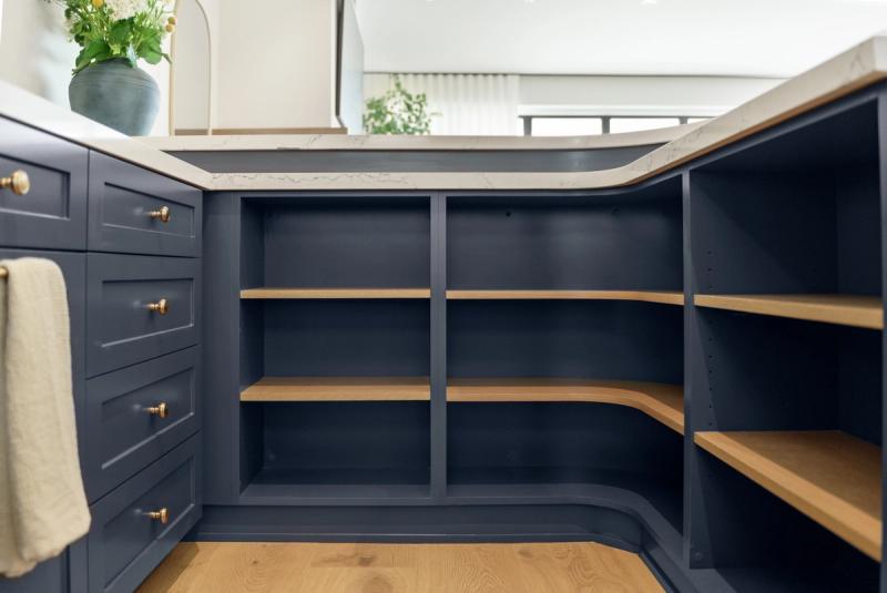 A navy blue kitchen island with open lower shelving featuring natural oak wood shelves, viewed straight on. The island has a white marble-style countertop and is part of a larger kitchen with matching navy cabinetry and brass hardware.