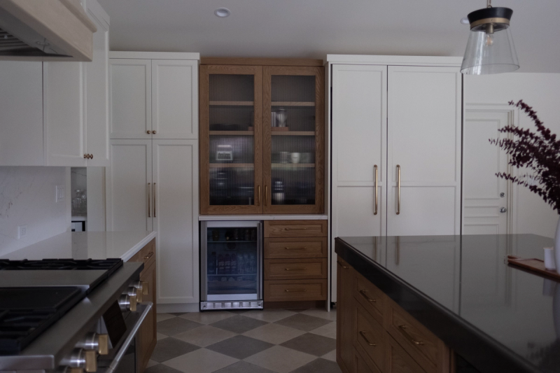 A transitional-style kitchen featuring cream shaker cabinets with brass hardware, a warm oak hutch with reeded glass doors and stacked drawers, a built-in wine cooler, checkered tile flooring, and a dark stone island countertop.