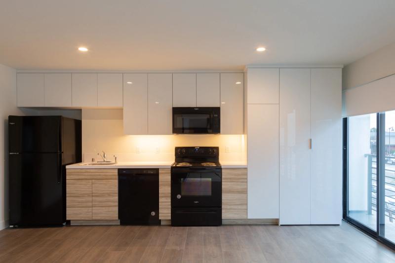 A modern kitchen featuring high-gloss white upper cabinets, light wood-grain lower cabinets, black Whirlpool appliances, and under-cabinet lighting, with a tall white pantry cabinet and sliding glass door to a balcony on the right.