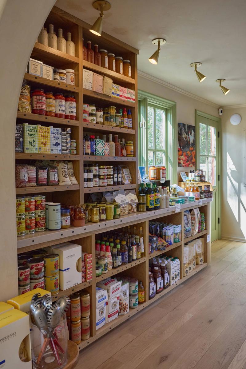 A warmly lit specialty food shop interior featuring floor-to-ceiling oak built-in shelving with an arched alcove, stocked with pantry goods, sauces, broths, and dry goods. The shelving runs along the wall beside green-trimmed windows with natural light flooding in.
