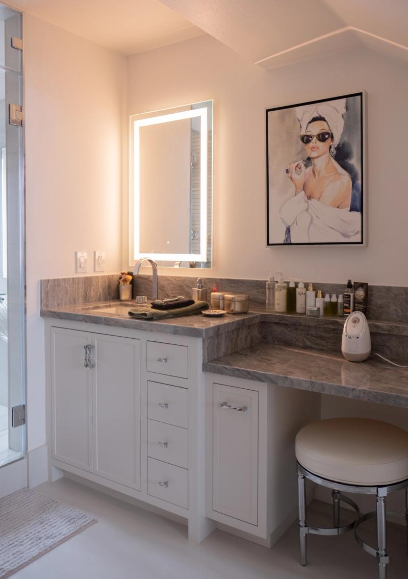 A bright, elegant bathroom vanity featuring white shaker-style cabinetry with chrome hardware, a marble-look stone countertop, an LED backlit mirror, and an adjacent makeup vanity area with a cushioned stool. Fashion artwork hangs on the wall above the vanity counter.