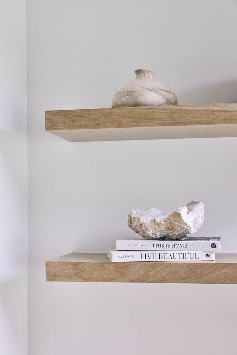 Two light wood floating shelves mounted on a white wall, styled with a ceramic vase on the upper shelf and stacked coffee table books topped with a raw crystal geode on the lower shelf.