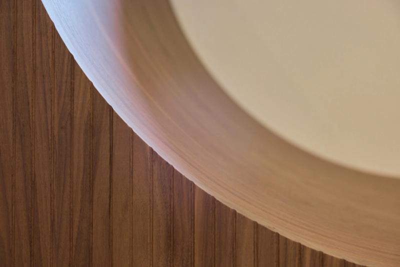 A close-up architectural detail showing a curved cream-colored ceiling element meeting a vertical walnut wood panel wall, with soft ambient backlighting illuminating the transition.