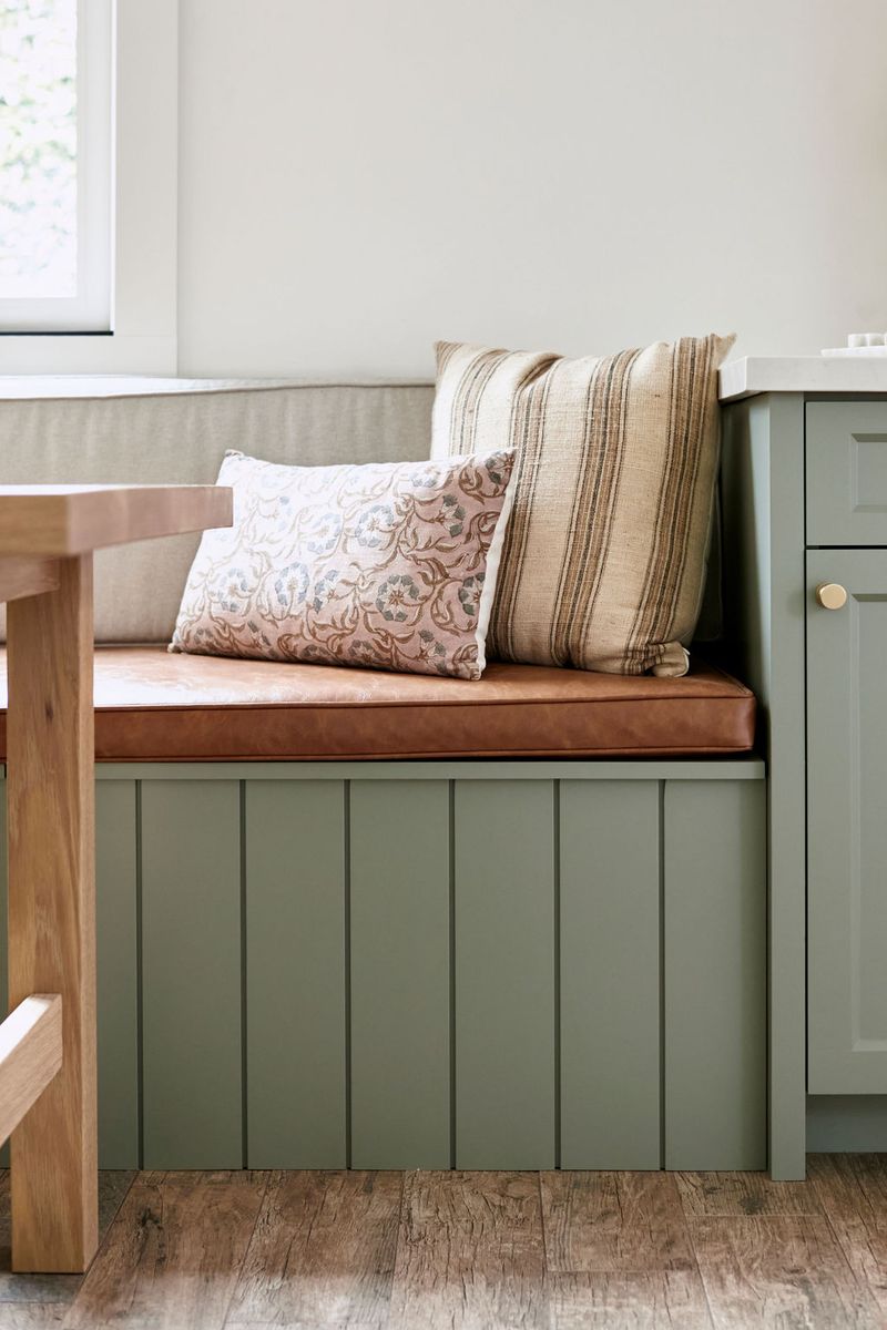 A built-in kitchen banquette with sage green shiplap paneling, a cognac leather seat cushion, and decorative throw pillows, positioned next to matching sage green cabinetry with brass hardware.