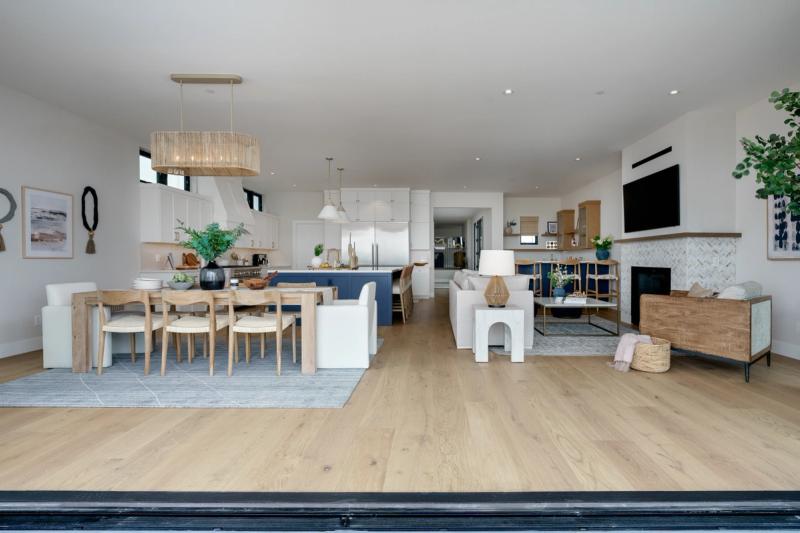 A bright, open-concept living space featuring white shaker cabinetry, a navy blue kitchen island, natural wood dining furniture, and a herringbone tile fireplace surround with light oak hardwood floors throughout.