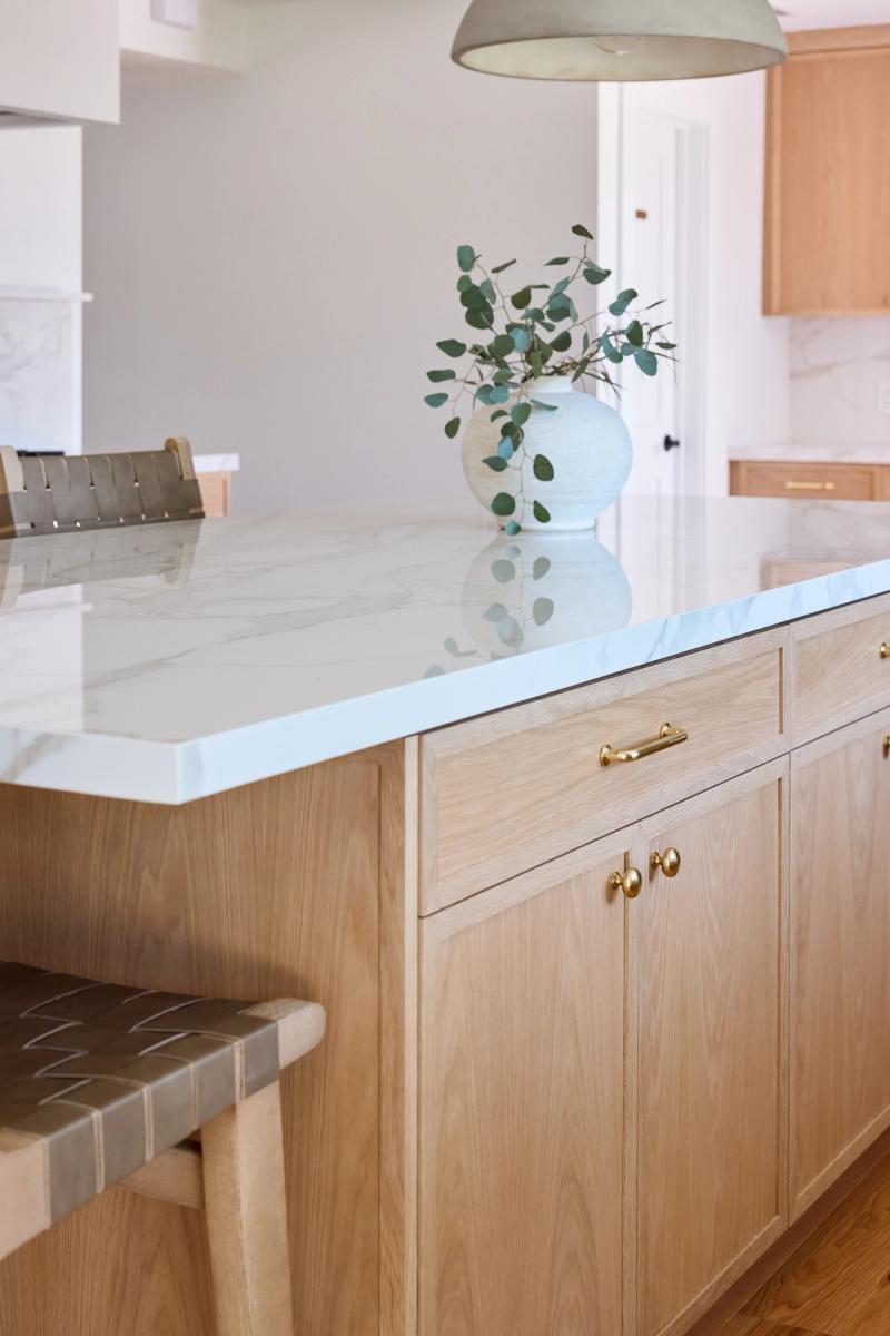A close-up of a custom white oak kitchen island featuring a polished white marble countertop, brass hardware, and a decorative white vase with eucalyptus branches. Leather woven barstools are tucked underneath.