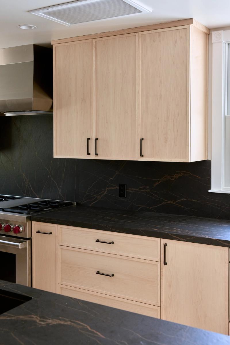 A modern kitchen featuring light maple shaker-style cabinets with matte black hardware, dark black-and-gold veined marble countertops and full-height backsplash, and a stainless steel professional range.
