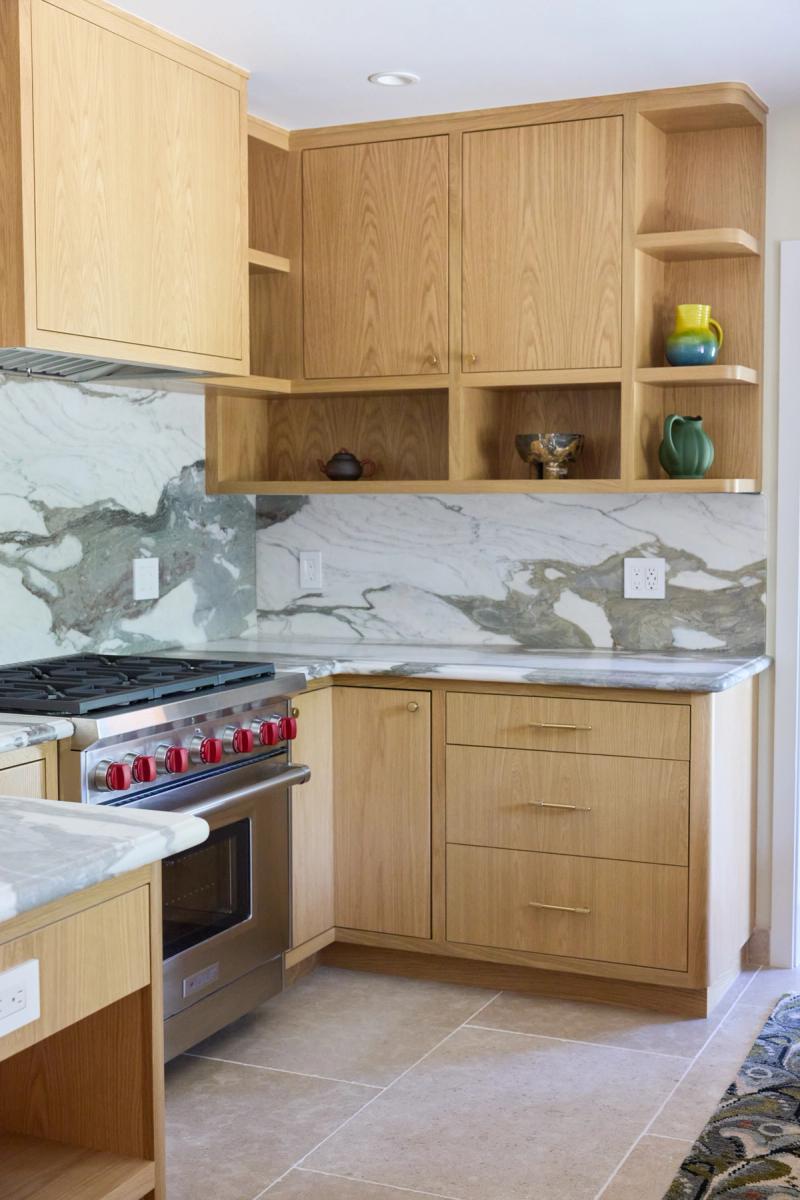 A modern kitchen featuring warm white oak cabinetry with brass hardware, dramatic marble slab backsplash and countertops, open display shelving, and a stainless steel range with red knobs.