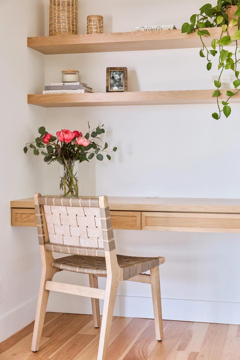 A built-in home office featuring a floating white oak desk with drawers and matching floating shelves styled with books, candles, woven baskets, a framed photo, and trailing plants. A woven leather chair sits at the desk with a vase of pink roses.