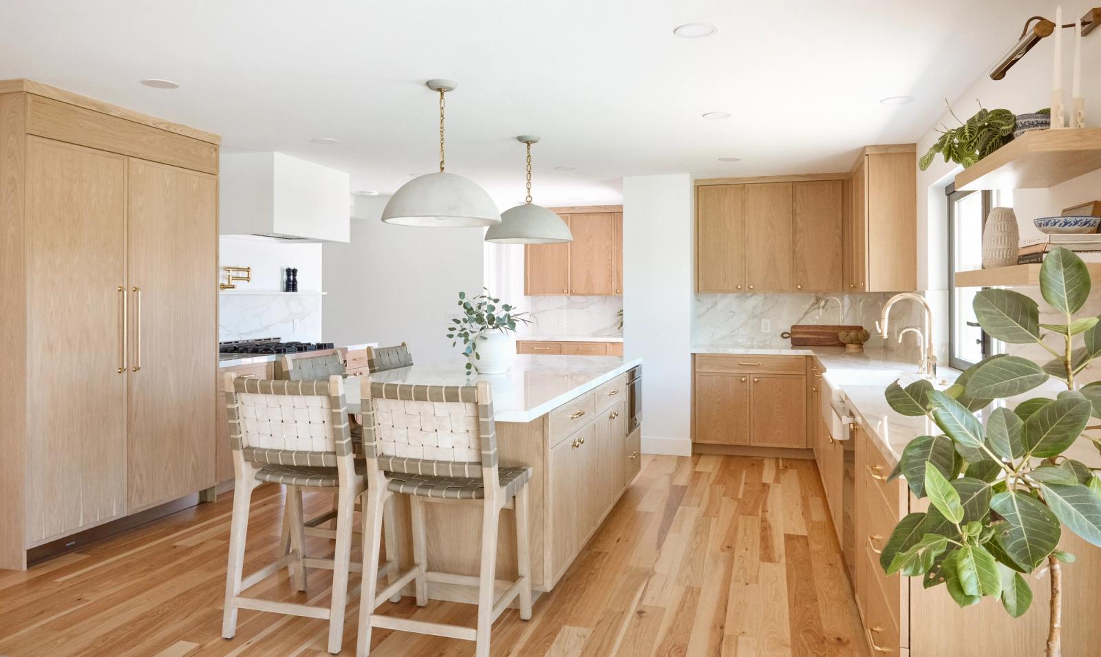 A bright, airy kitchen featuring natural white oak cabinetry, a large center island with woven leather bar stools, marble backsplash, brass hardware, and sage green dome pendant lights.