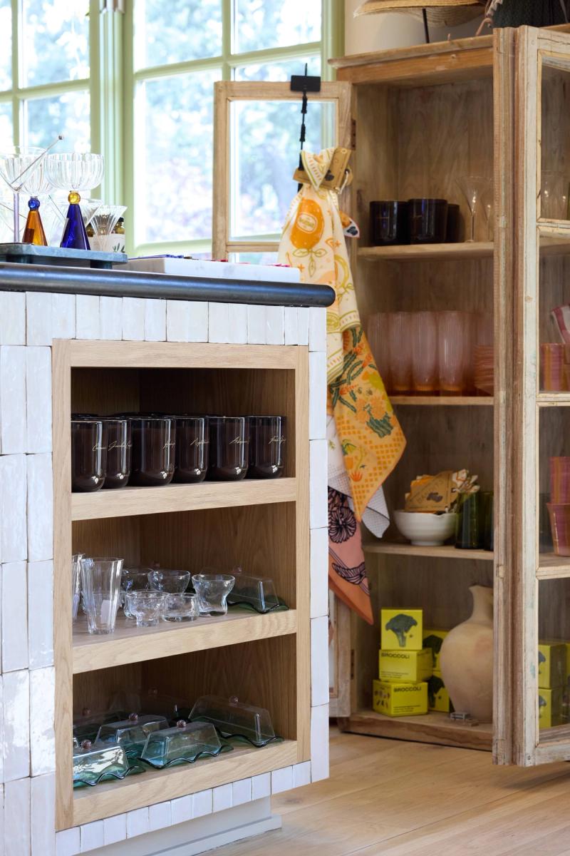 A retail or kitchen display area featuring a white zellige-tiled island with natural oak open shelving holding glassware, beside a rustic wood cabinet displaying colorful homewares and broccoli-branded boxes.