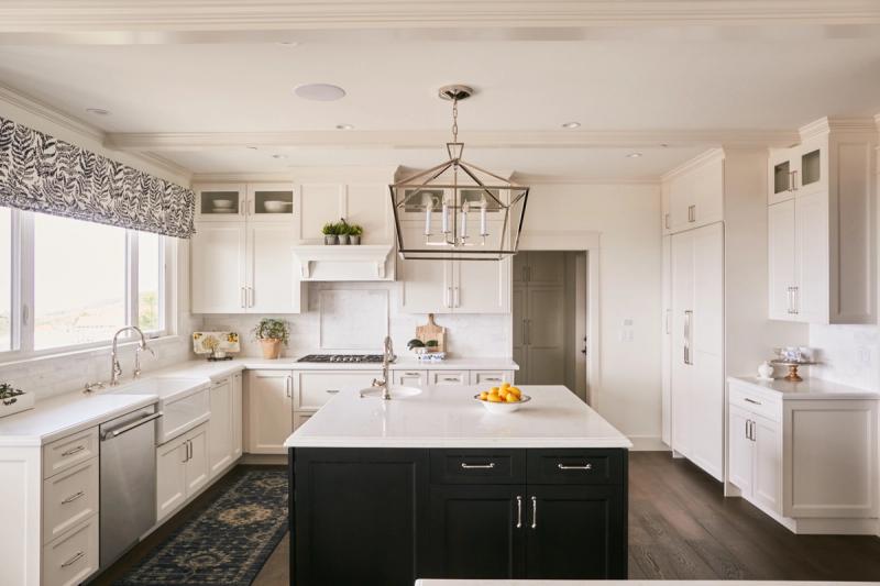 A bright, transitional-style kitchen featuring white shaker cabinets, a dark navy island with white quartz countertop, geometric pendant light, and dark hardwood floors.