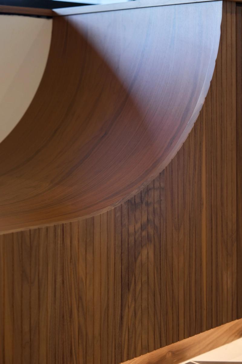 A close-up detail shot of a curved walnut wood panel with a sweeping circular cutout, showcasing fine grain and custom craftsmanship in a built-in millwork piece.