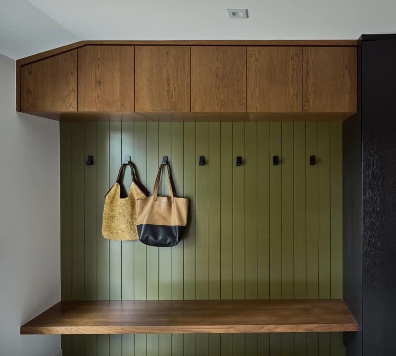 A custom mudroom built-in featuring olive green vertical shiplap paneling with black metal coat hooks, a floating walnut bench, and warm wood upper cabinets. Two bags hang from the hooks.