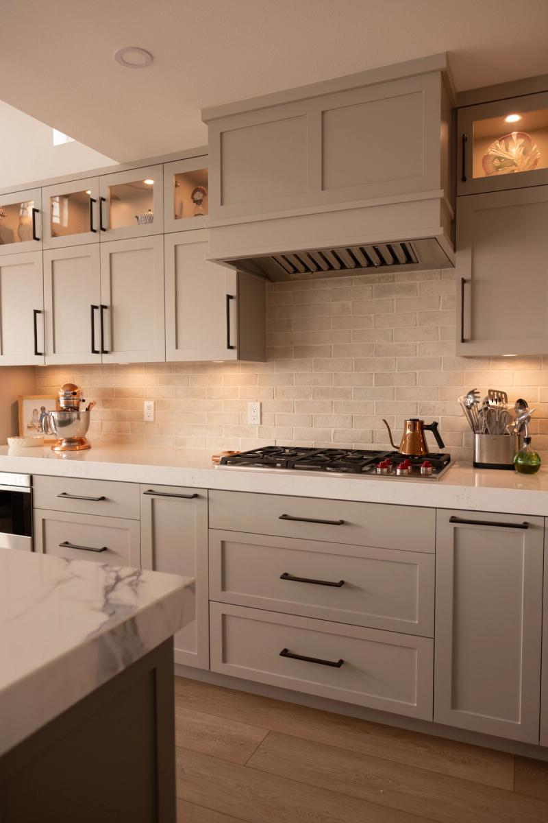 A beautifully designed kitchen featuring greige shaker-style cabinetry with matte black hardware, a custom built-in range hood, cream brick backsplash, white quartz countertops, and glass-front upper cabinets with interior lighting.