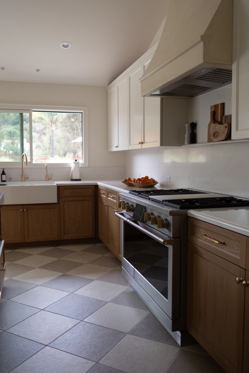 A bright, transitional-style kitchen featuring warm oak lower cabinets with brass hardware, white upper cabinets, a farmhouse sink, professional stainless steel gas range with brass knobs, and a checkerboard tile floor.
