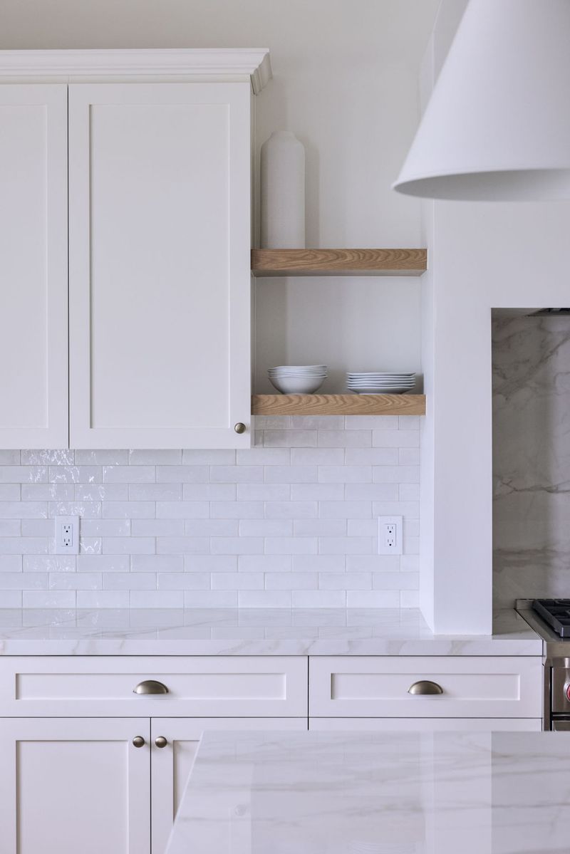 A bright, all-white custom kitchen featuring shaker-style cabinetry, marble countertops, white subway tile backsplash, brushed nickel hardware, and floating oak open shelves styled with white dishes.