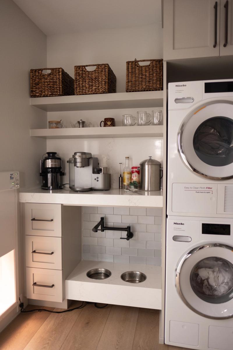 A built-in coffee bar with open white shelving, wicker storage baskets, and a coffee appliance station beside a stacked Miele washer and dryer. A lower built-in pet feeding station with two stainless steel bowls and a pot filler faucet is tucked beneath the counter.