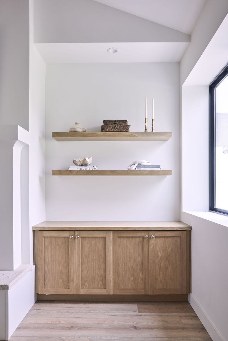 A recessed niche in a bright white room features two floating white oak shelves styled with decorative objects and books, above a four-door white oak base cabinet with a light wood countertop.