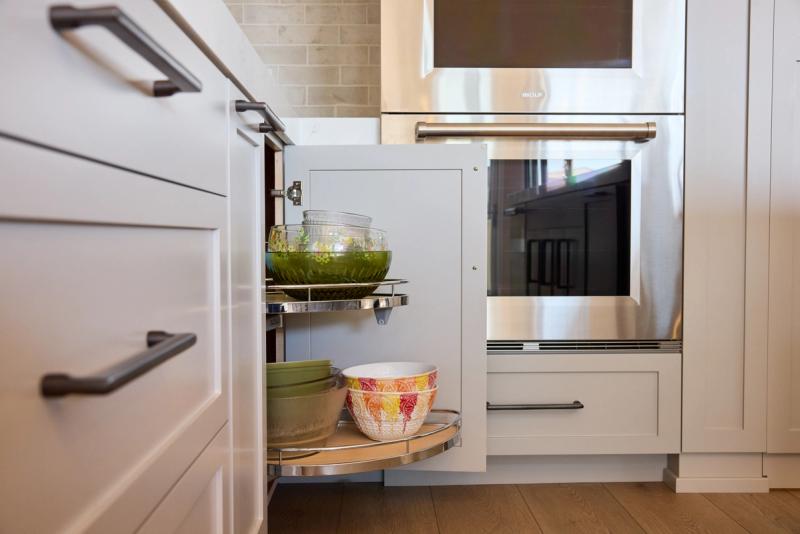 An open corner base cabinet with a pull-out lazy Susan organizer holding colorful mixing bowls and glass dishes, flanked by white shaker-style cabinetry and a stainless steel Wolf wall oven.