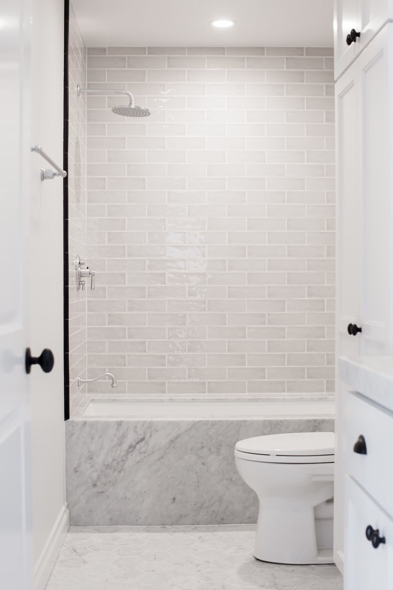 A bright, elegant bathroom featuring a built-in bathtub with Carrara marble surround, white subway tile walls, a rainfall showerhead, and white cabinetry with matte black hardware.