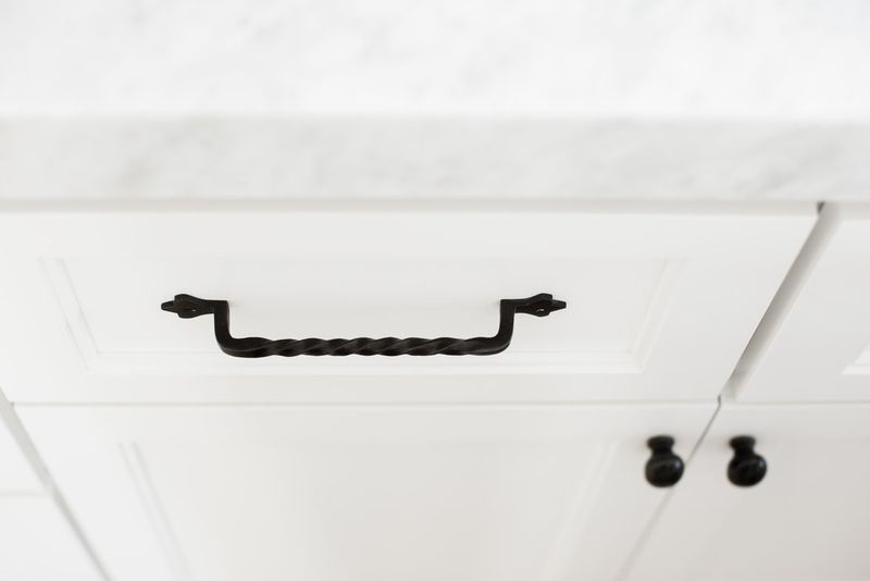 A close-up of a white bathroom vanity drawer featuring a twisted black iron pull handle, with a marble countertop above and black cabinet knobs visible to the right.