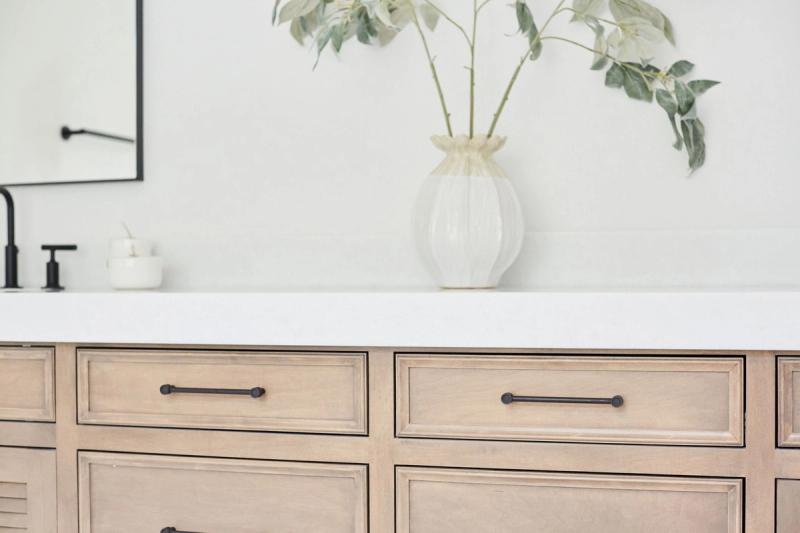 A close-up of a custom bathroom vanity featuring light natural wood cabinetry with raised panel drawer fronts, matte black bar pulls, and a clean white quartz countertop topped with a ribbed white ceramic vase holding green eucalyptus stems.