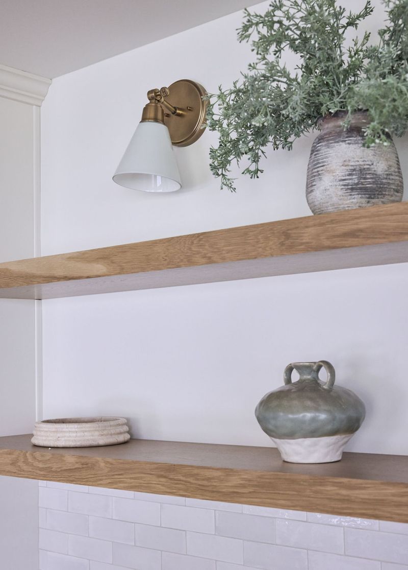 Two floating white oak shelves mounted on a white wall, styled with ceramic vases and greenery, with a brass and white wall sconce above. White subway tile is visible at the bottom of the frame.