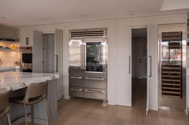 A high-end transitional kitchen featuring floor-to-ceiling white shaker cabinetry surrounding a stainless steel Sub-Zero French door refrigerator with glass doors and a built-in wine cooler column. A marble waterfall island with bar stools is visible in the foreground.
