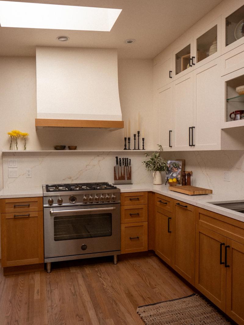 A custom kitchen featuring warm honey-toned lower cabinets and cream upper cabinets with black hardware, a stainless steel professional range beneath a painted wood range hood, marble-look quartz countertops and backsplash, and a skylight above.