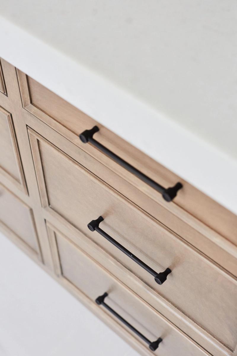 Close-up of custom light wood bathroom vanity drawers with raised panel detailing and matte black bar pulls, topped with a white stone countertop.