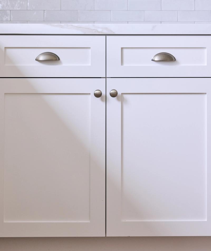 Close-up of white shaker-style base cabinets featuring two drawers with cup pulls and two lower cabinet doors with round knobs, topped with a marble countertop and white subway tile backsplash.