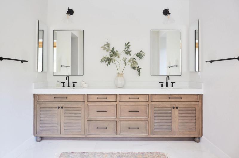 A wide double bathroom vanity featuring light wood cabinetry with louvered cabinet doors, matte black hardware, white quartz countertop, dual undermount sinks, and two rectangular black-framed mirrors with wall sconces above.
