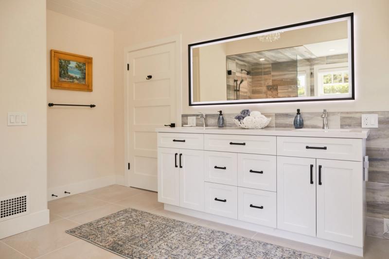 A bright master bathroom featuring a large white shaker-style double vanity with black hardware, marble-look countertops, and a wide backlit mirror. The room includes light tile flooring, a patterned area rug, and a framed landscape painting on the wall.
